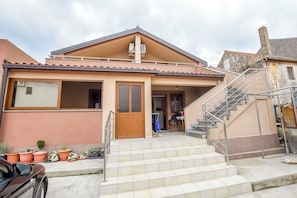 Un edificio de dos pisos con fachada color crema, una puerta de madera y una escalera que sube al nivel superior.