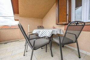 Un pequeño balcón exterior con una mesa y dos sillas, una ventana con contraventanas de madera y suelo de baldosas.