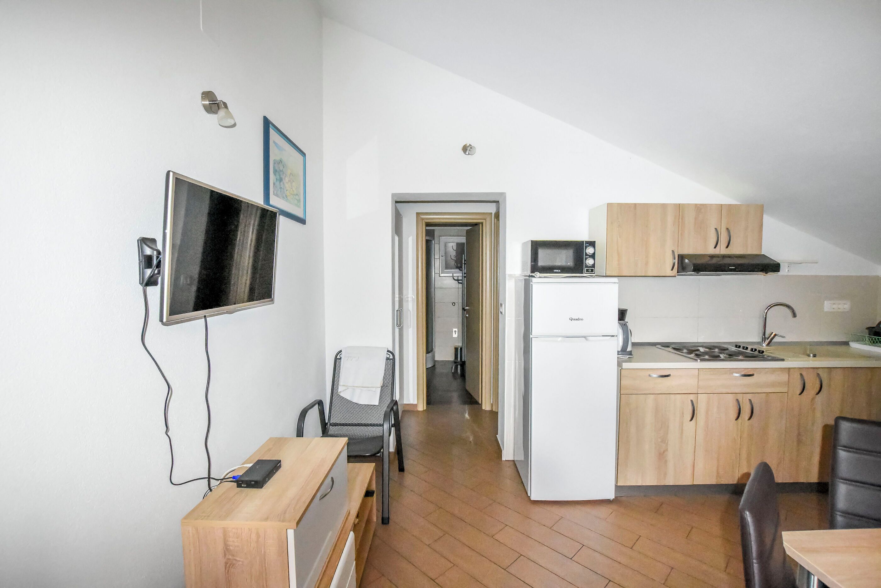 A compact kitchen with a refrigerator, microwave, and wooden cabinets. A dining area with a table and chairs is visible, and a television is mounted on the wall.