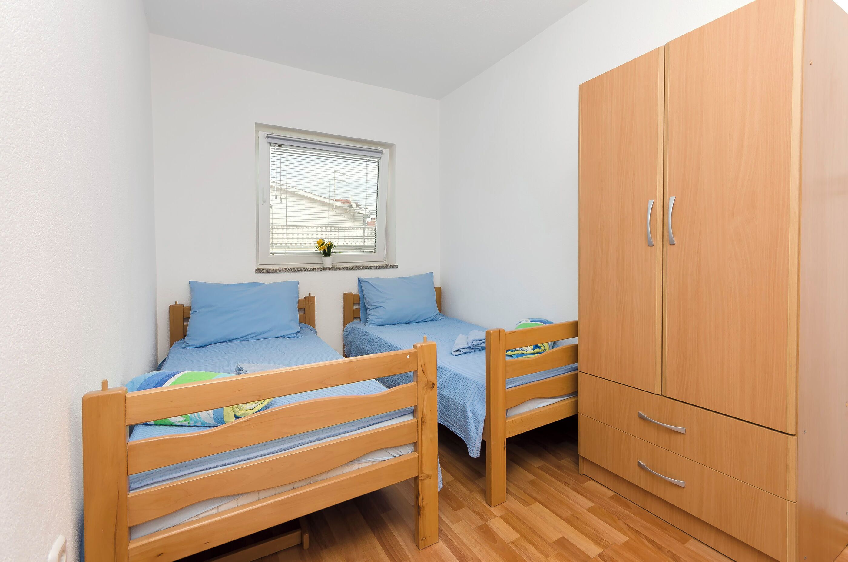 A small, tidy room with two wooden bunk beds, a wooden wardrobe, and a window with blinds.
