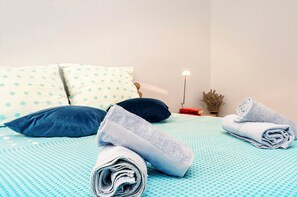 Cama bem arrumada com colcha azul texturizada, duas almofadas e uma toalha enrolada.