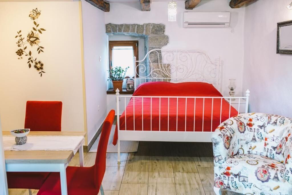 A bedroom with a white metal bed frame, red bedding, a red chair, a table with a bowl, and a wall with a floral design.