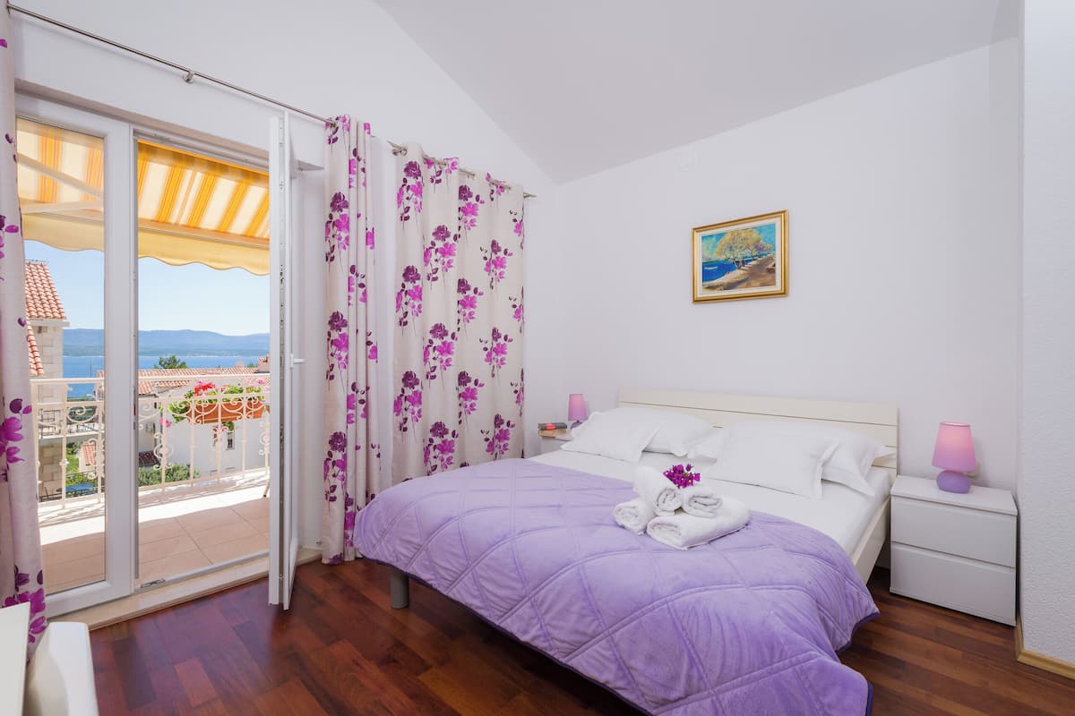 Una camera da letto con un letto, un comodino, una lampada, un quadro e vista sul mare attraverso una porta aperta.