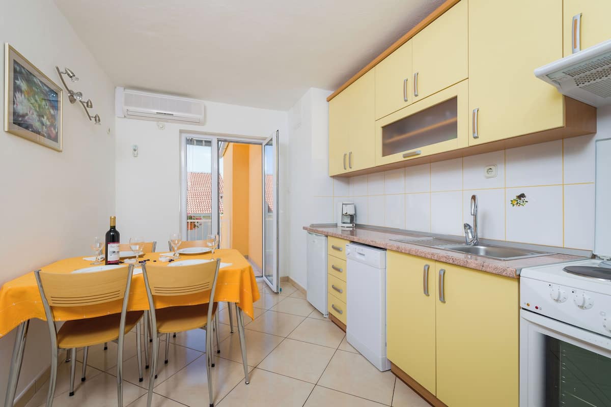 Cucina con mobili gialli, lavastoviglie bianca e fornello. Si nota un tavolo da pranzo con sedie e una bottiglia di vino.