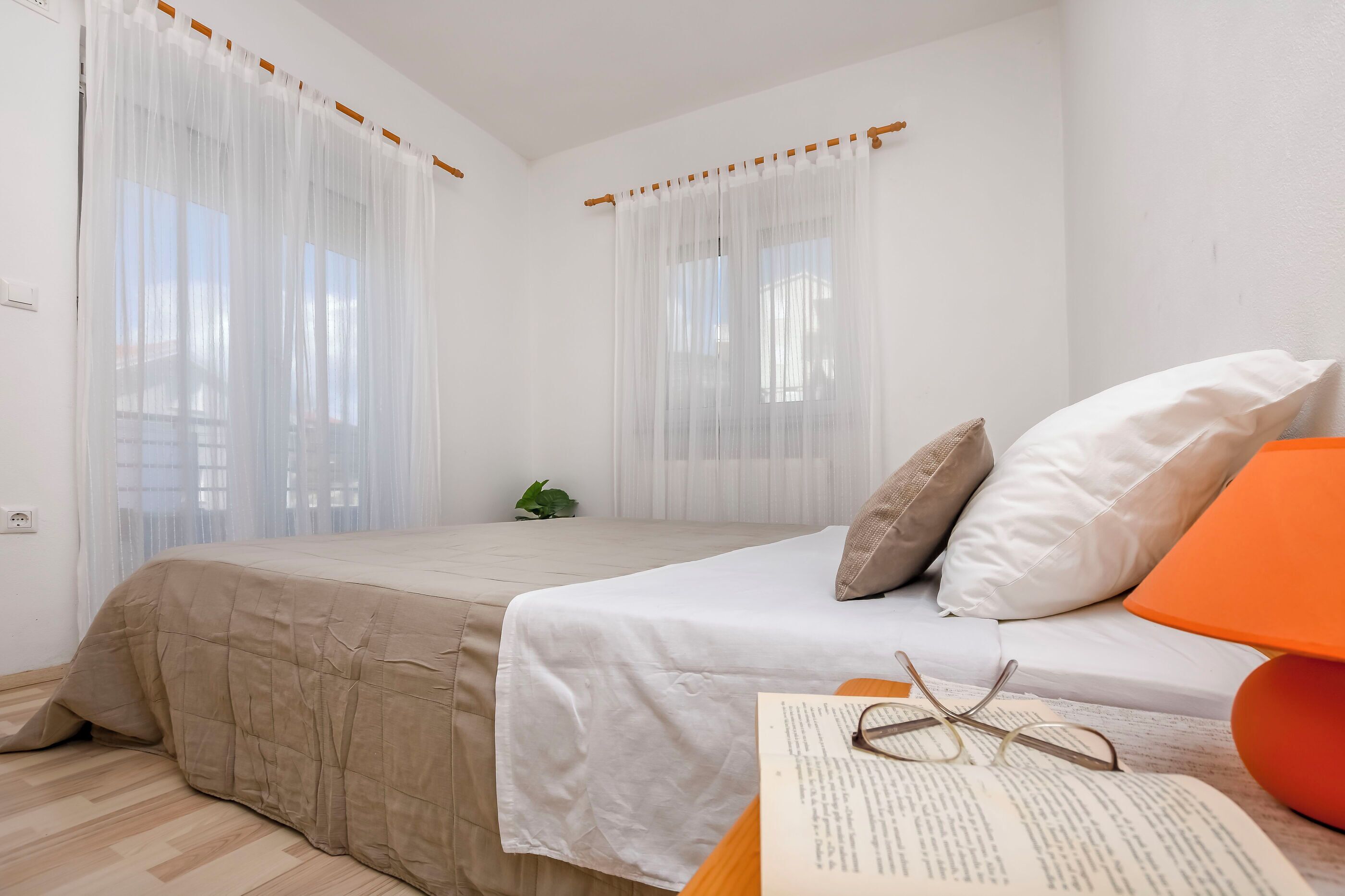 Une chambre avec deux lits, du linge de lit blanc, une petite table de chevet avec un livre et des lunettes, et une plante.