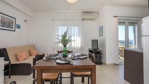 A dining area with a table set for four, a sofa with cushions, a green vase with a plant, and a view of the outdoors through a window. - Apartment Prcela 2 (Primosten)