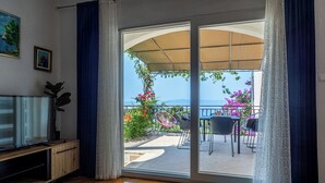 A room with a sliding glass door leading to an outdoor patio with a table and chairs, a TV on a wooden cabinet, and a potted plant. - Apartment Vito (Podgora)