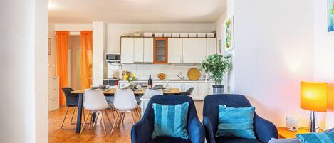 A modern living room with a dining area, featuring a sofa set, a coffee table with flowers, and a kitchen in the background.