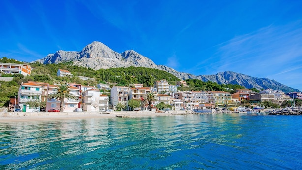A coastal town with buildings along the waterfront, clear blue water, and mountains in the background. - Apartment Vito (Podgora)