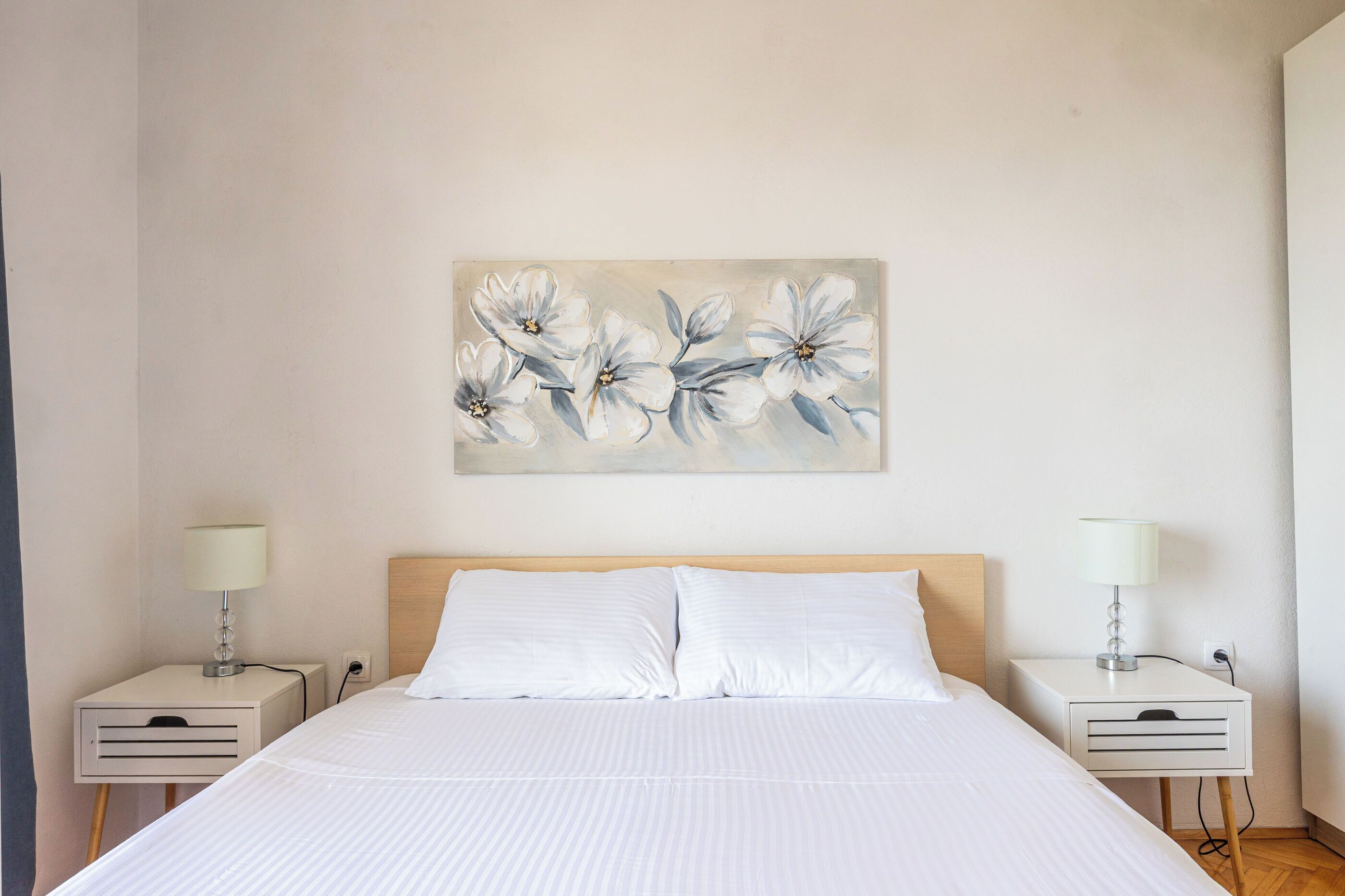 A neatly made bed with white linens, two bedside tables with lamps, and a floral painting on the wall.