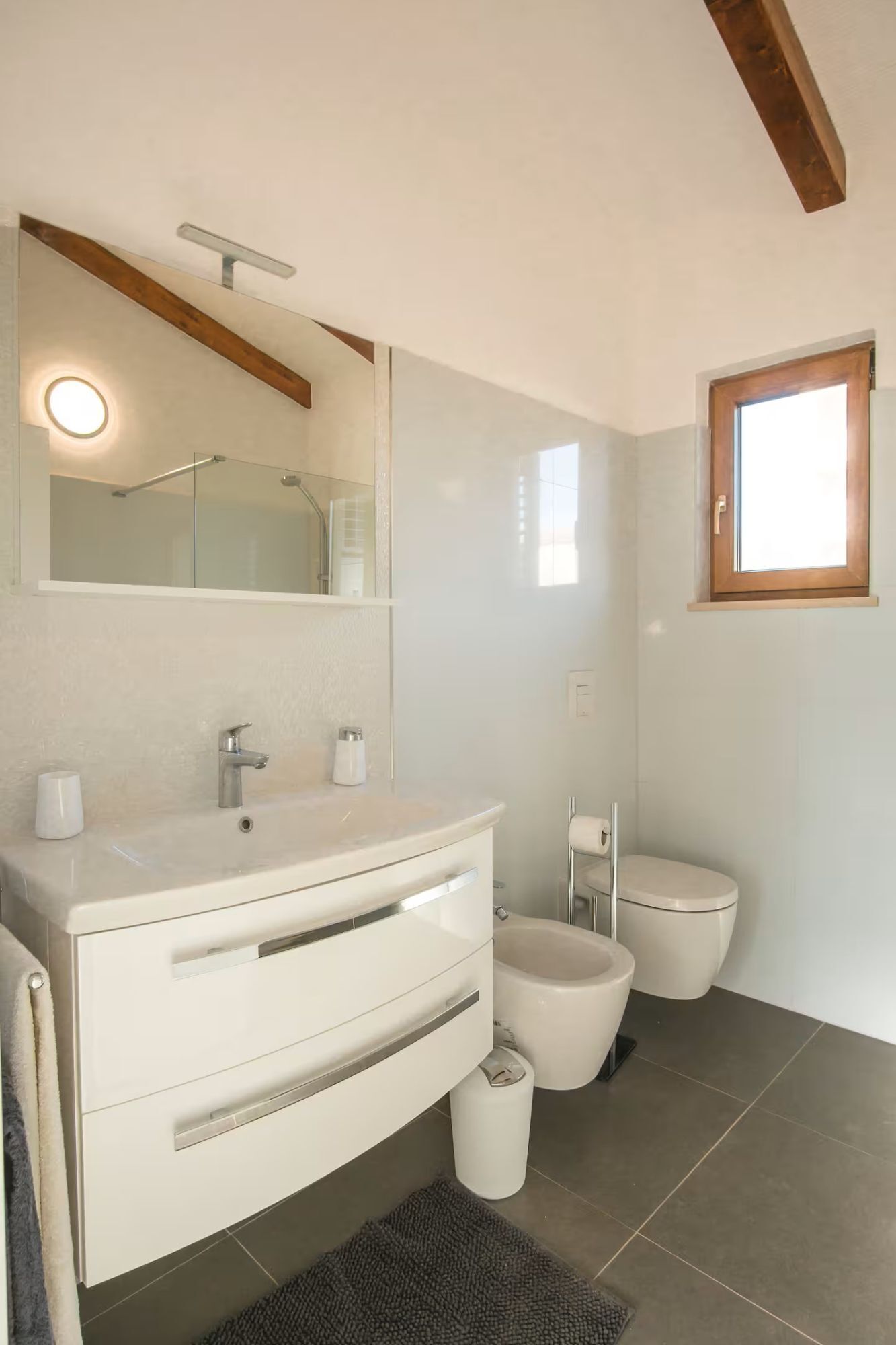 Et moderne badeværelse med hvid kommode, glasbrusekabine, toilet og et vindue.