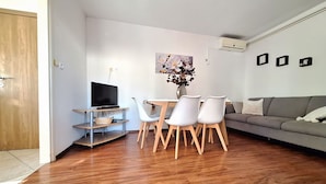 Ein modernes Wohnzimmer mit Holzboden, einem grauen Sofa, einem Esstisch mit weißen Stühlen, einem Flachbildfernseher und einer Vase mit Blumen.