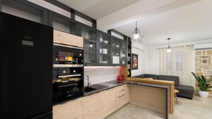A modern kitchen with a black refrigerator, wooden cabinets, and a built-in oven. There is a dining area with a grey sofa and a potted plant. - Villa Lipa Dora (Bol)