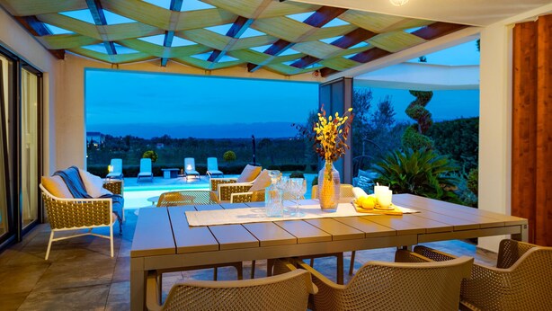 A dining area with a table set for four, surrounded by wicker chairs, a vase with flowers, and a view of the landscape. - Villa Royal Green Three (Policnik)