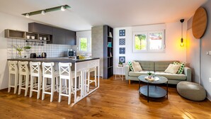 A modern kitchen with a central island, white bar stools, a dining table, and a living area with a sofa and a round coffee table. - Villa Royal Green Three (Policnik)