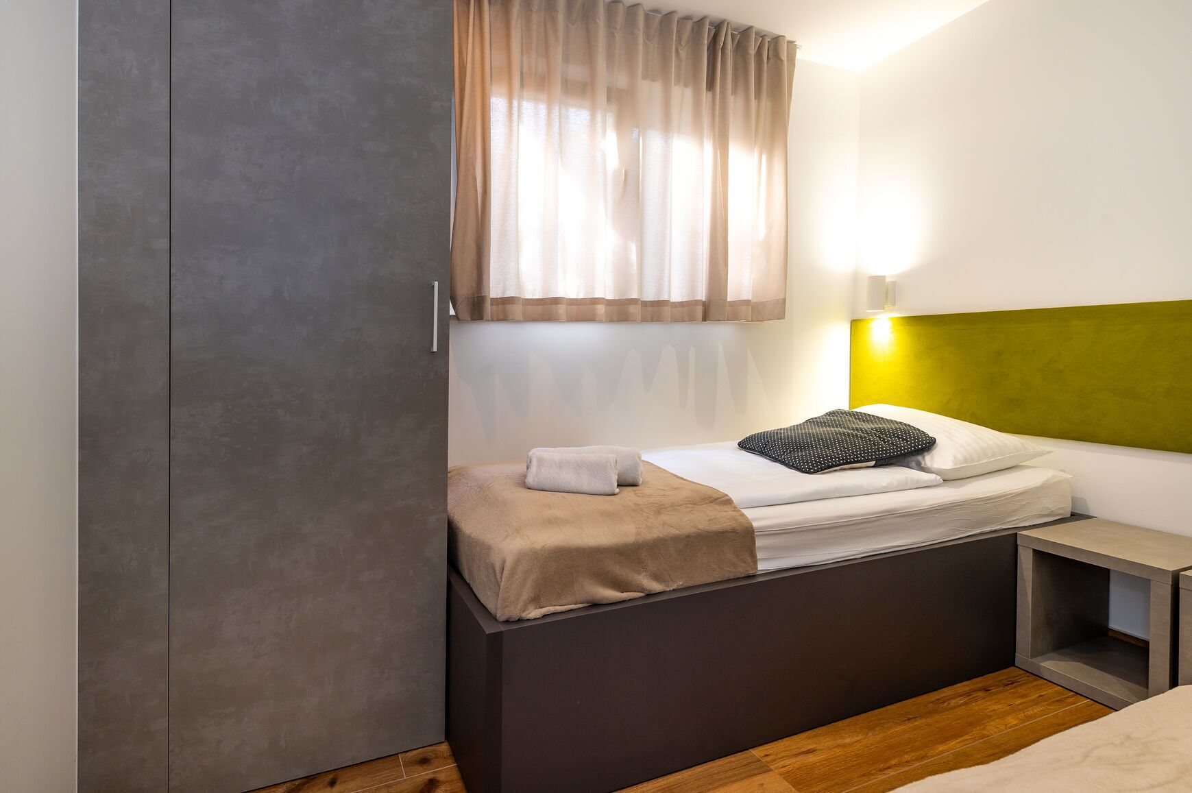 A modern bedroom with a bed, a bedside table, a wardrobe, and a window with sheer curtains.