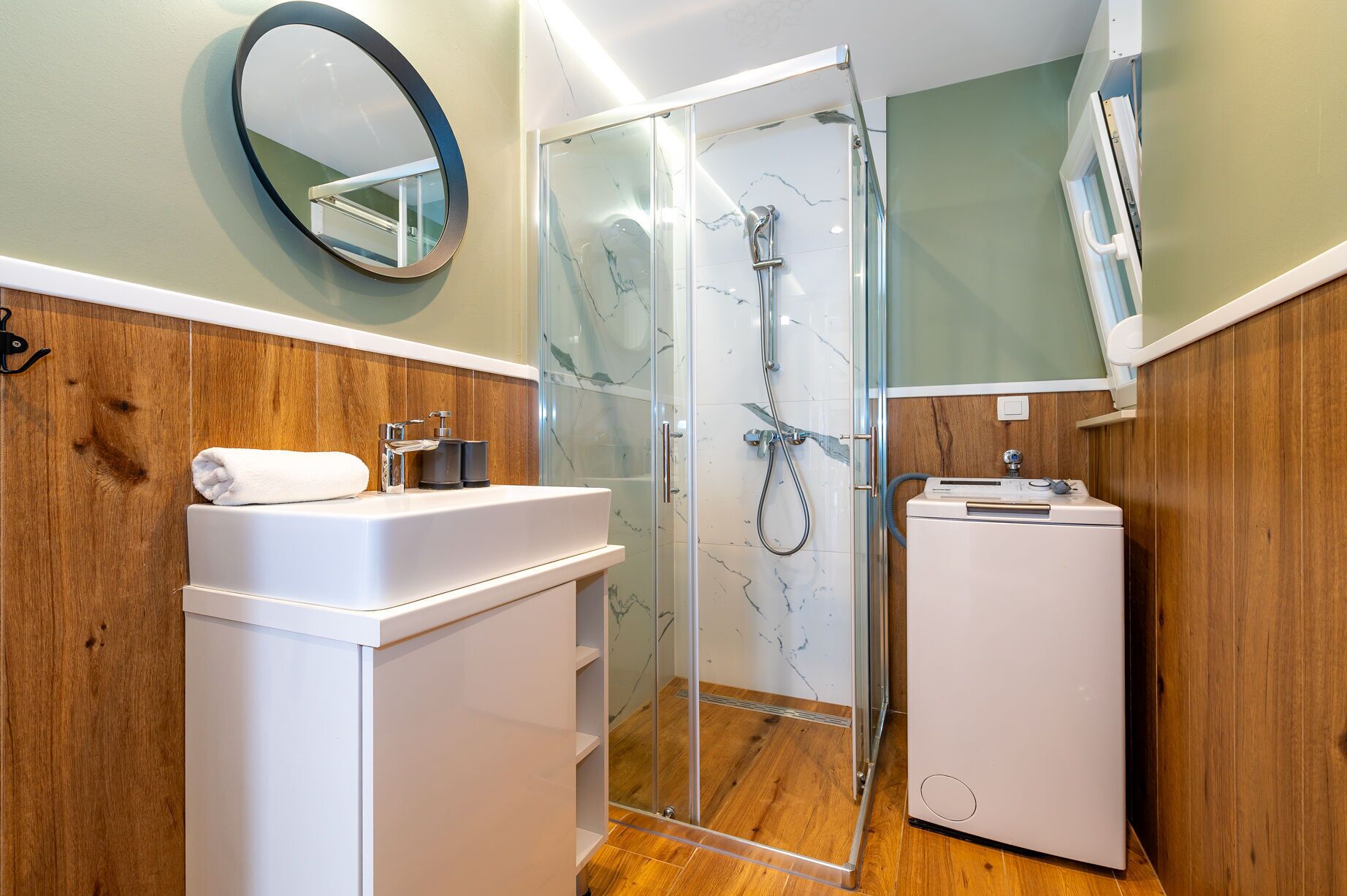 A modern bathroom with a glass-enclosed shower, a white sink with a towel, and a compact washing machine.