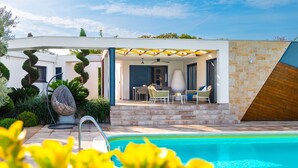 A modern outdoor pool area with a covered seating area, a pool, and surrounding greenery. - Villa Royal Green Three (Policnik)