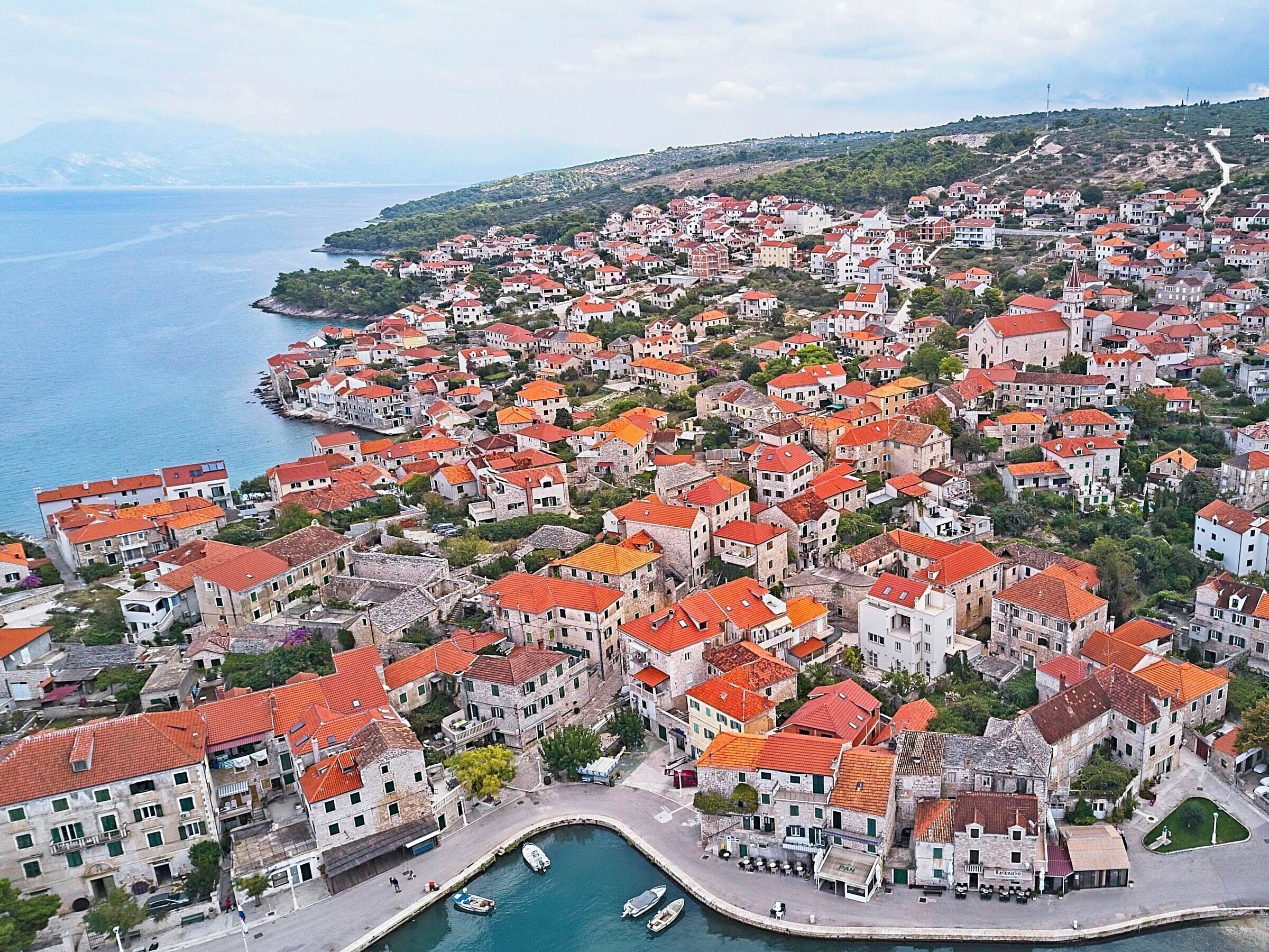 Une ville côtière aux bâtiments aux toits rouges, dotée d’un port de plaisance et bordée de collines.