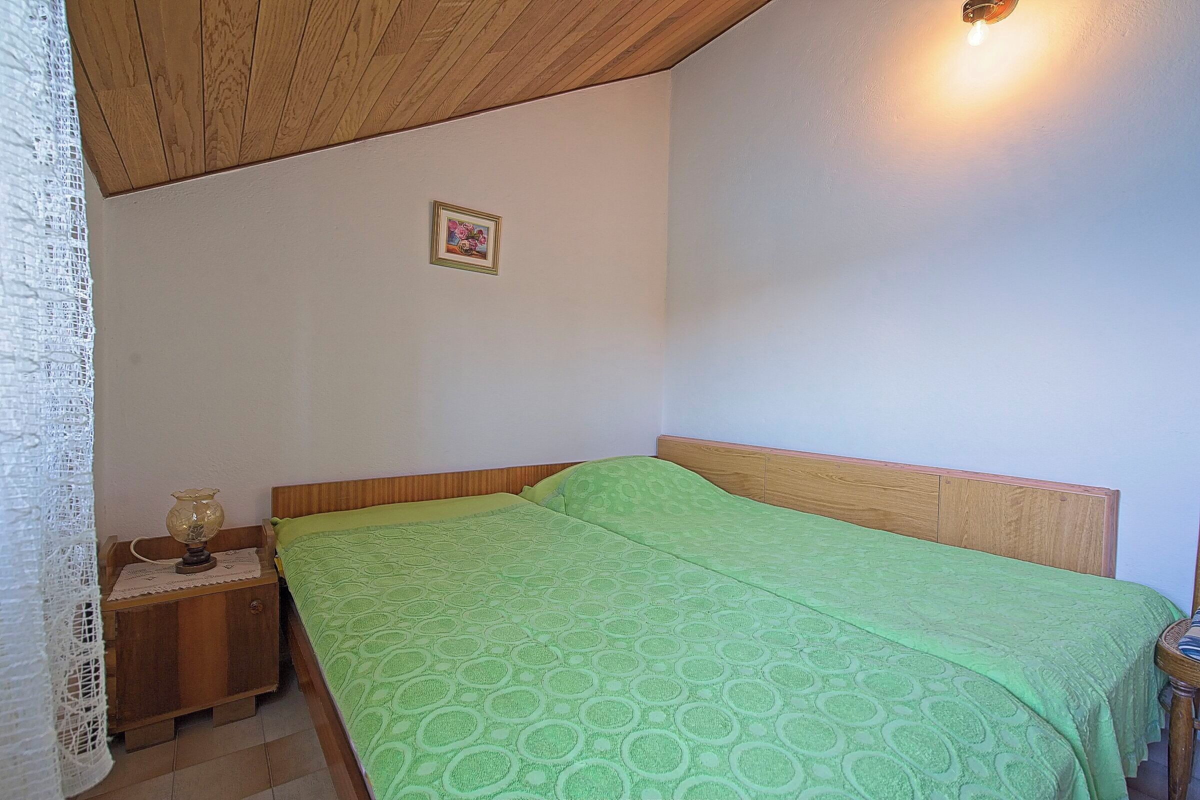 A bedroom with a wooden headboard, a green patterned bedspread, a bedside table with a lamp, and a small framed picture on the wall.
