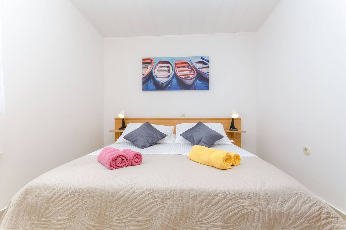 Un lit bien fait, avec une tête de lit matelassée, deux oreillers et des serviettes roulées, de couleur rose et jaune.