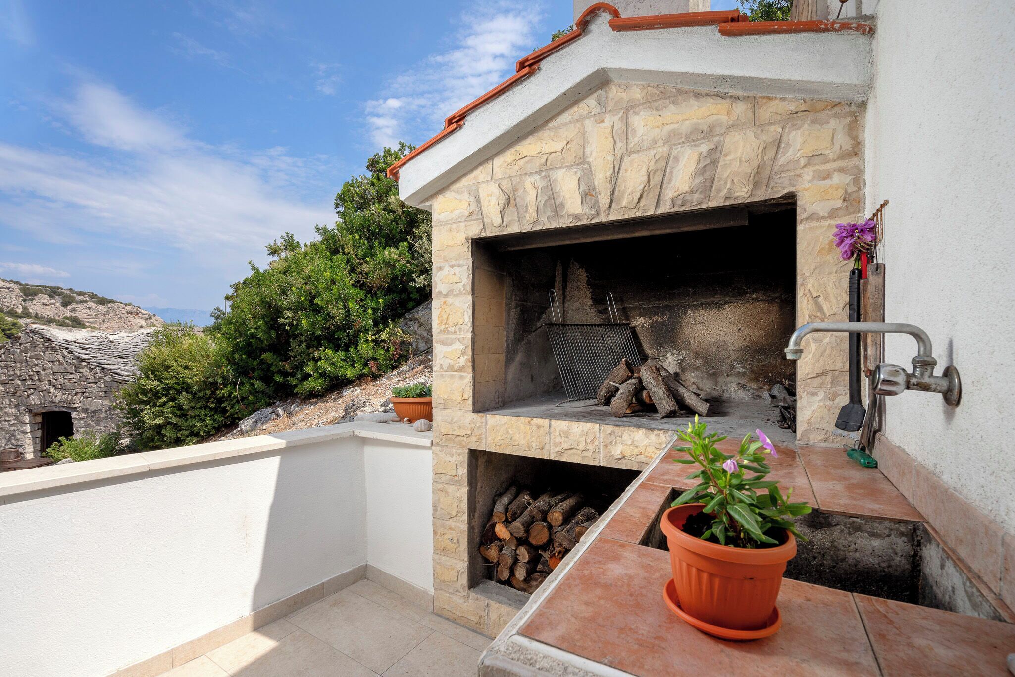 An outdoor stone fireplace with a metal grill, surrounded by a white wall and a potted plant.