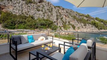 Una terrazza con un divano, sedie e un tavolo con bevande, affacciata su una costa rocciosa e un tratto d'acqua.