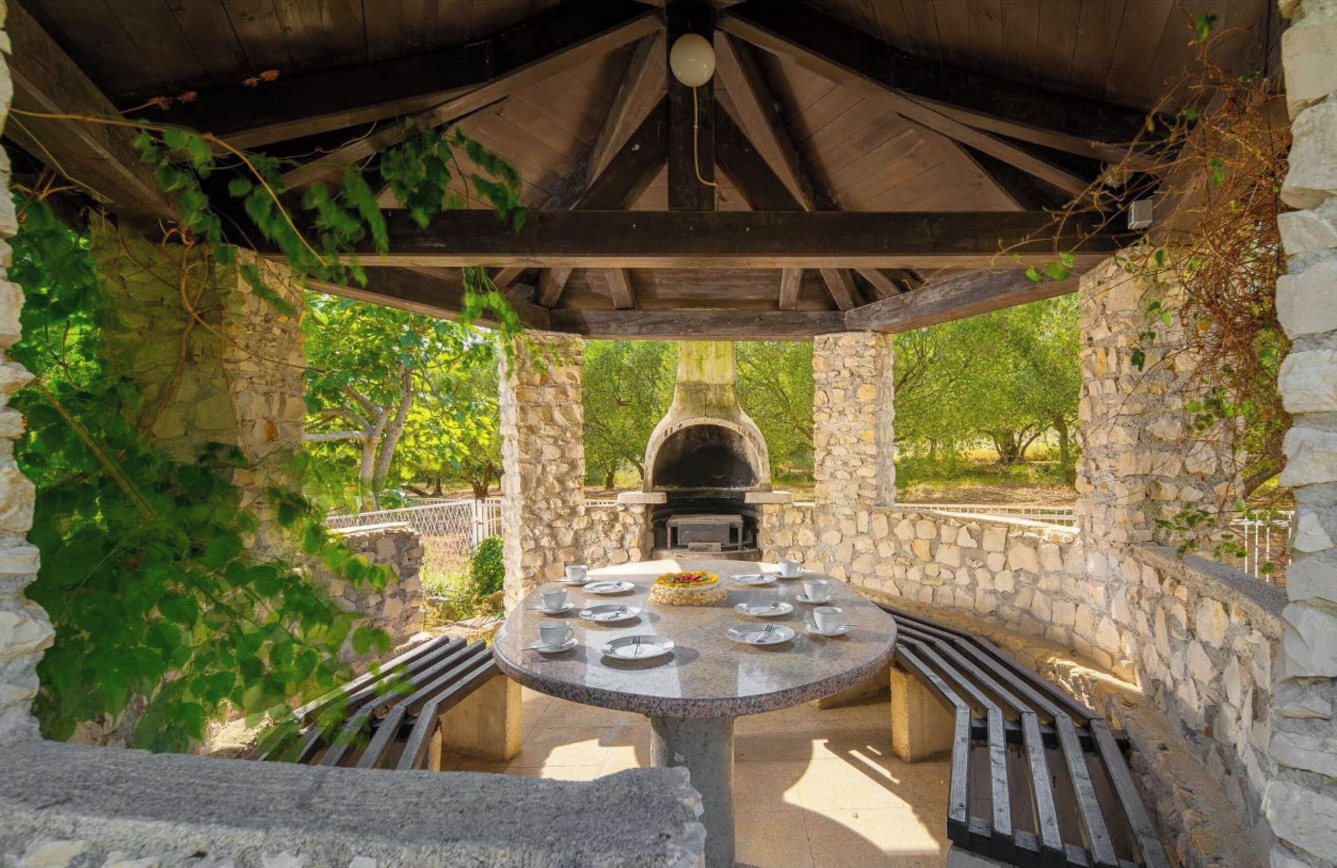Un área de estar al aire libre con arcos de piedra, una mesa redonda dispuesta para comer, rodeada de muros de piedra y vegetación.