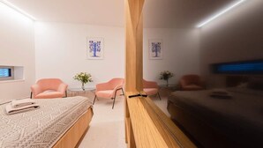 Une chambre d’hôtel moderne avec un grand lit, deux fauteuils roses, une petite table sur laquelle est posé un vase de fleurs, et des œuvres d’art accrochées aux murs.
