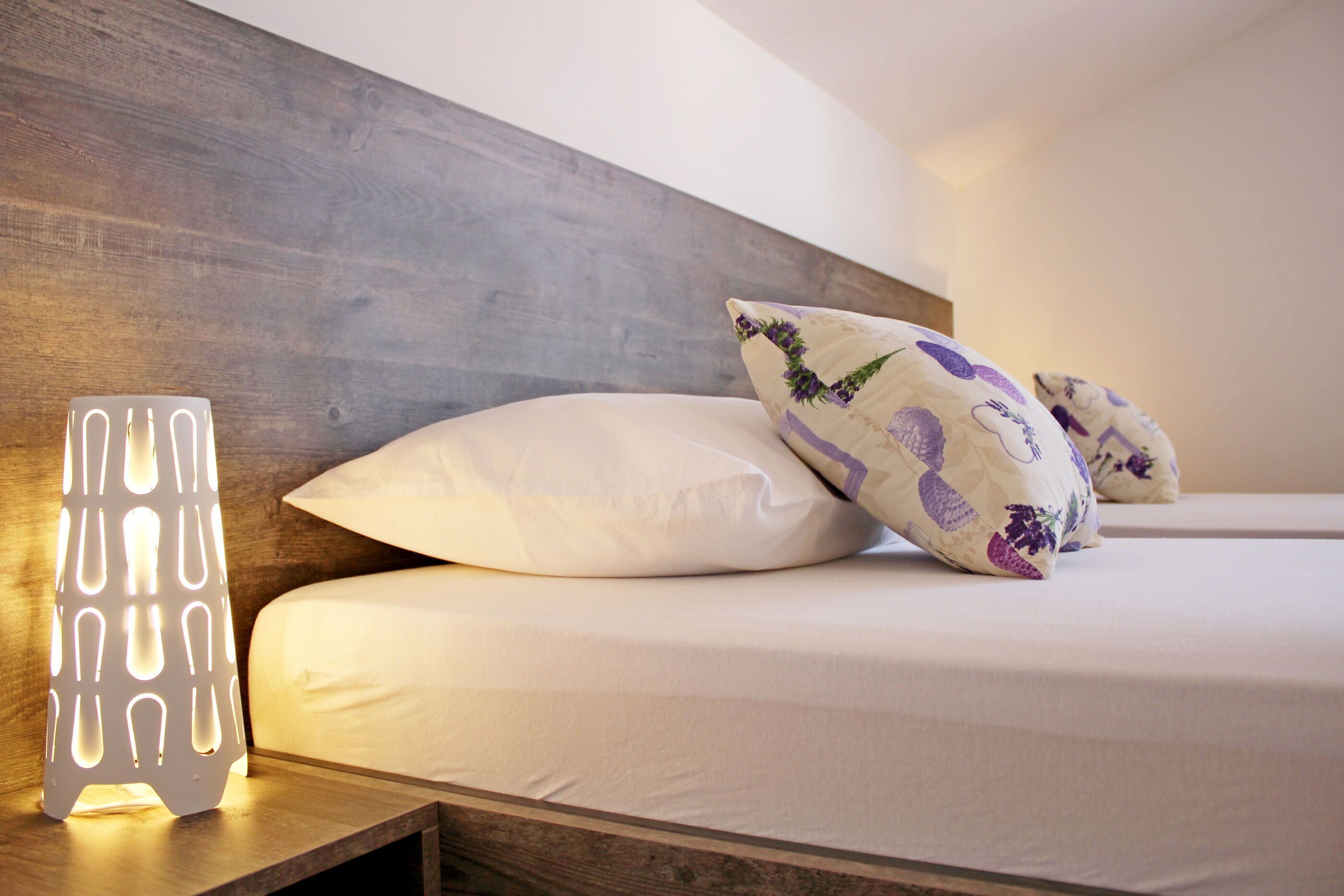 A neatly made bed with white linens and decorative pillows, a modern wooden headboard, and a wall-mounted light fixture.