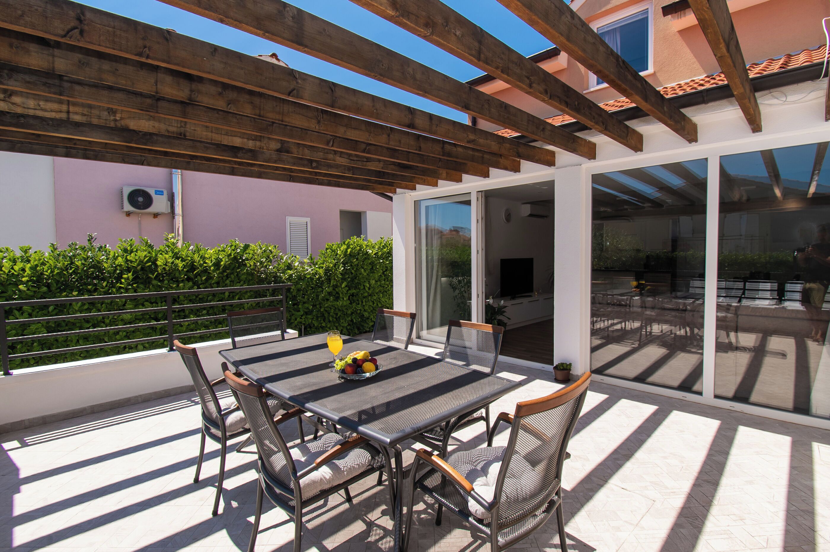 An outdoor patio with a table and chairs, a wooden pergola, and a glass door leading to a covered area.