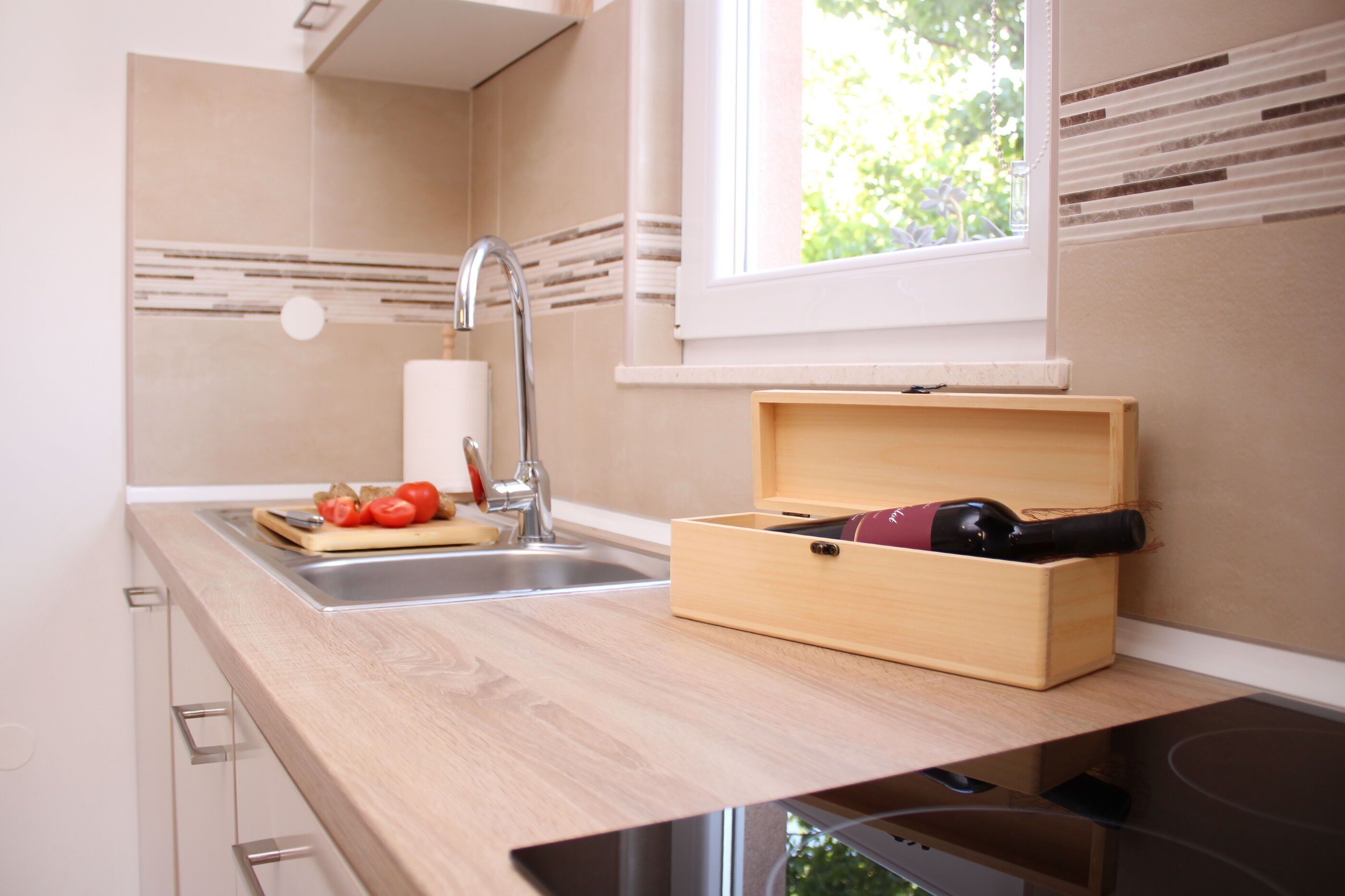 A kitchen with a wooden countertop, a stainless steel sink, a window with blinds, and a wine box with bottles.
