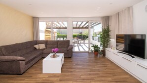 A modern living room with a brown sectional sofa, a white coffee table with flowers, a flat-screen TV, and a balcony with outdoor furniture. - Villa Divine (Vodice)