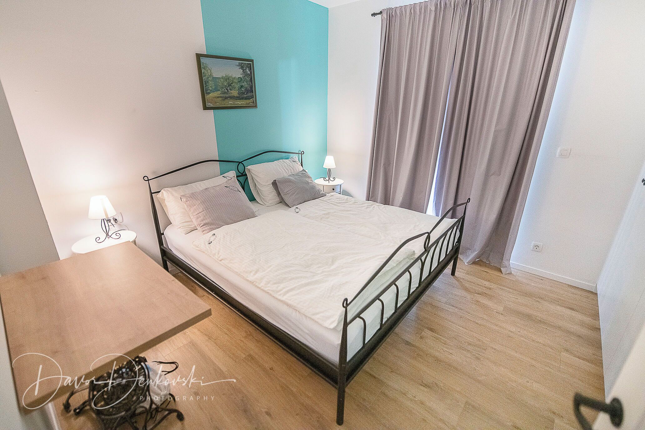 A neatly made bed with a metal frame, two bedside tables with lamps, a wooden desk, and a painting on the wall.