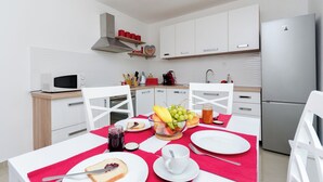 A modern kitchen with white cabinets, a stainless steel refrigerator, and a dining table set with plates, a bowl of fruit, and a red tablecloth. - Apartment Ivona (Vinodol)