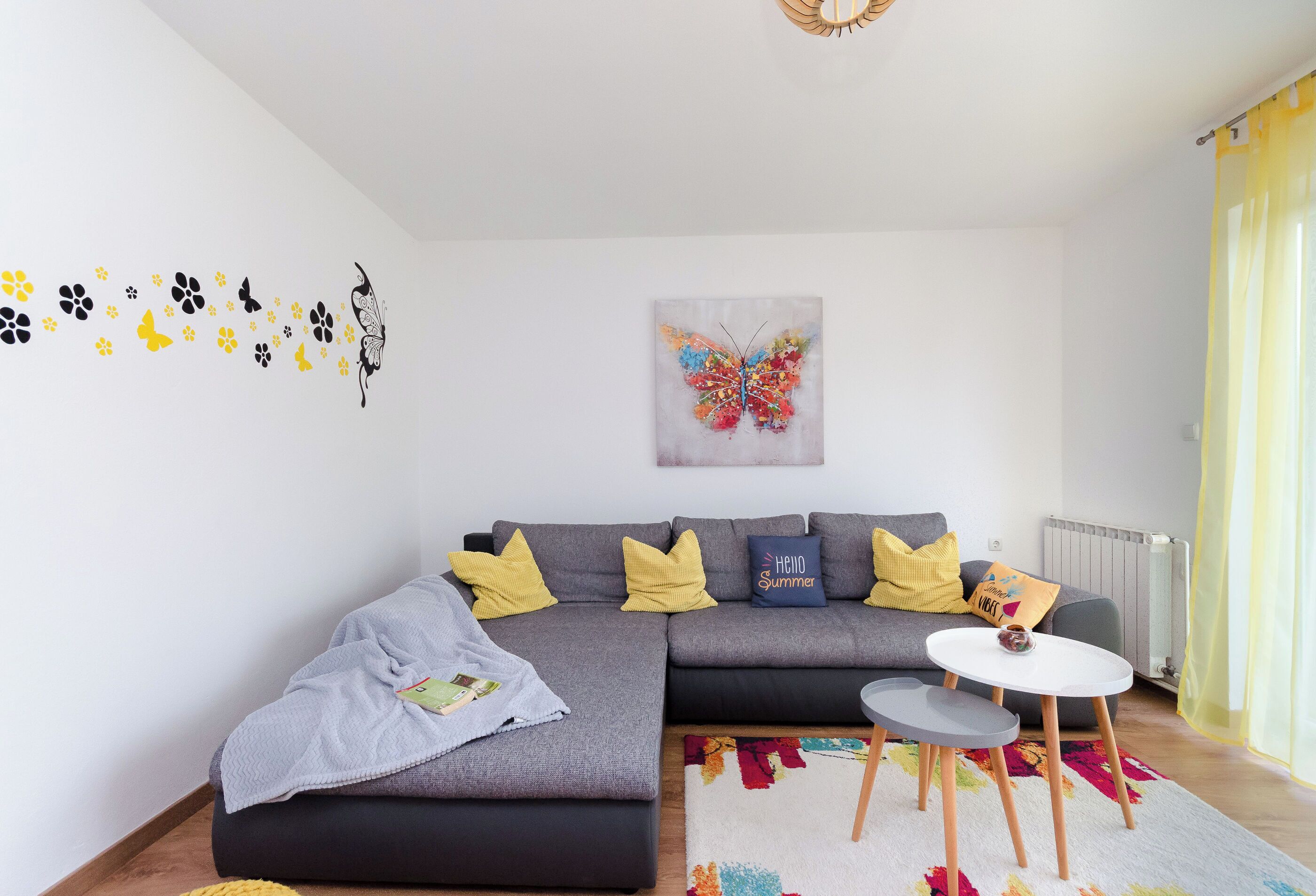 Ein modernes Wohnzimmer mit einem grauen Ecksofa, gelb-schwarzen Wandstickern, einem farbenfrohen Schmetterlingsbild und einem kleinen weißen Tisch.