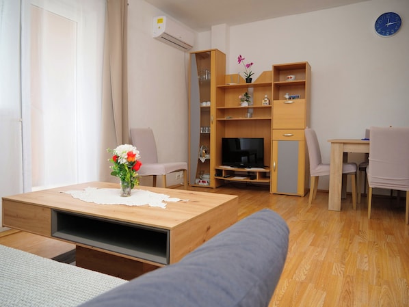 Een moderne woonkamer met een bank, een salontafel met een bloevas, een tv op een houten standaard en een eethoek met stoelen en een tafel.