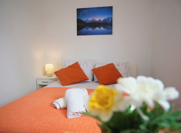 Un lit bien fait, avec une literie orange, deux oreillers, un oreiller blanc orné de broderies florales, une lampe de chevet et, au mur, un tableau représentant un paysage de montagne.