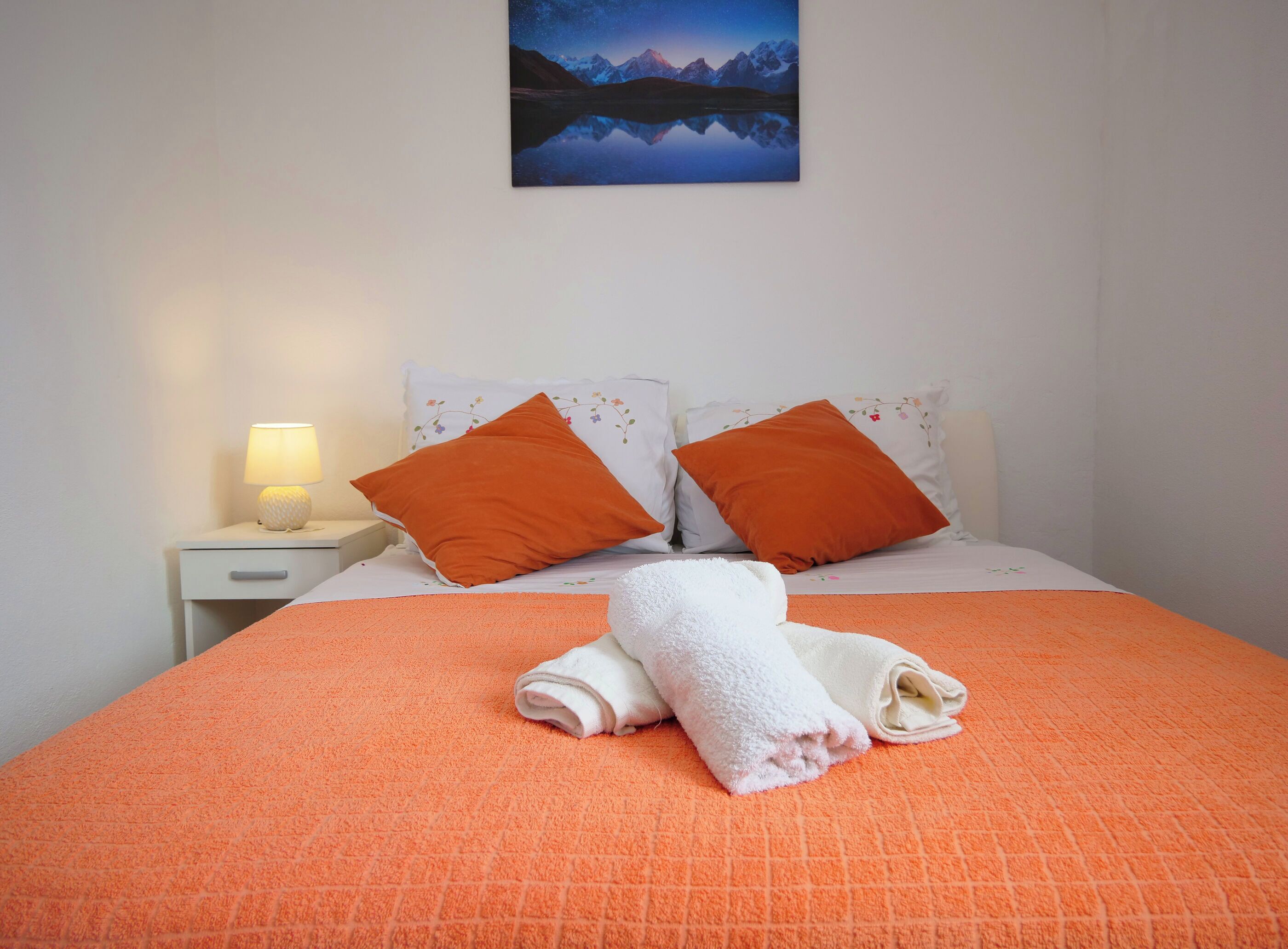 Un lit bien fait, recouvert d’une couverture orange, avec deux oreillers orange et une serviette blanche pliée posée sur le lit.