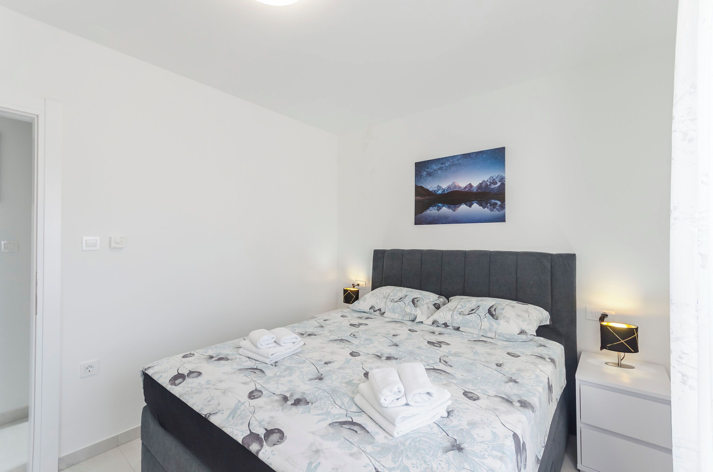 A bedroom with a bed, two bedside tables, a nightstand, a wall-mounted picture, and a door.