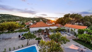 A villa with a swimming pool, a barbecue area, and a view of the sunset over a hilly landscape.