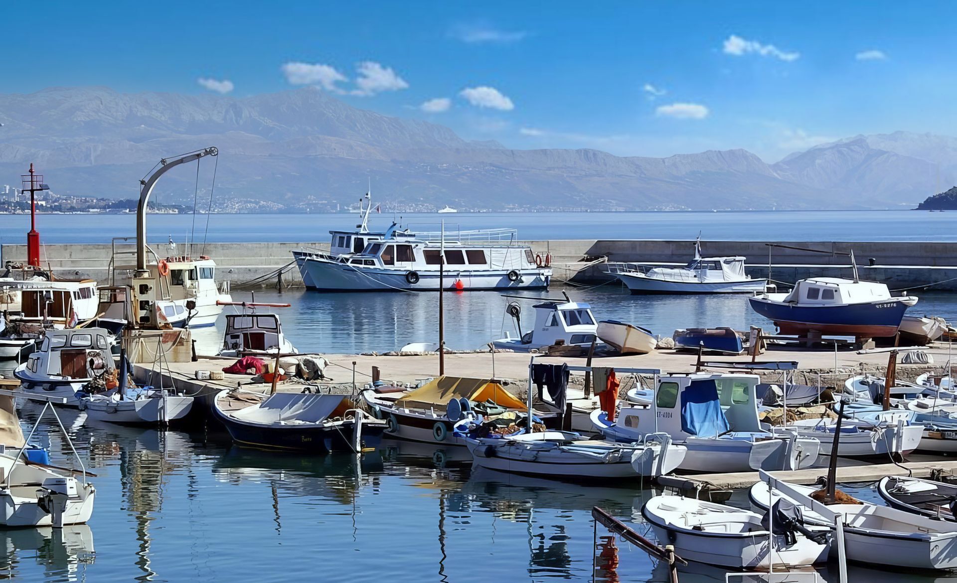 Una marina con varias embarcaciones amarradas, un faro al fondo y montañas a lo lejos.