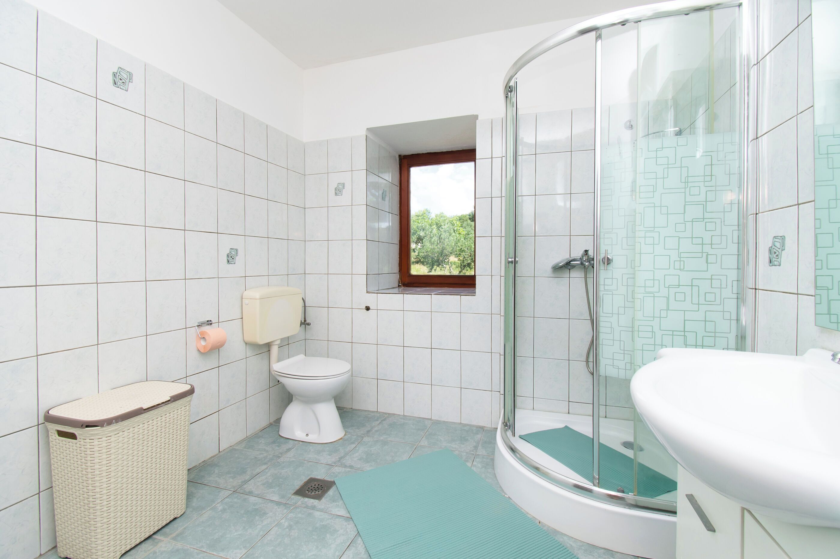 Ein Badezimmer mit Toilette, einer Dusche mit Glastür, einem Waschbecken, einem Fenster und einem Korb.