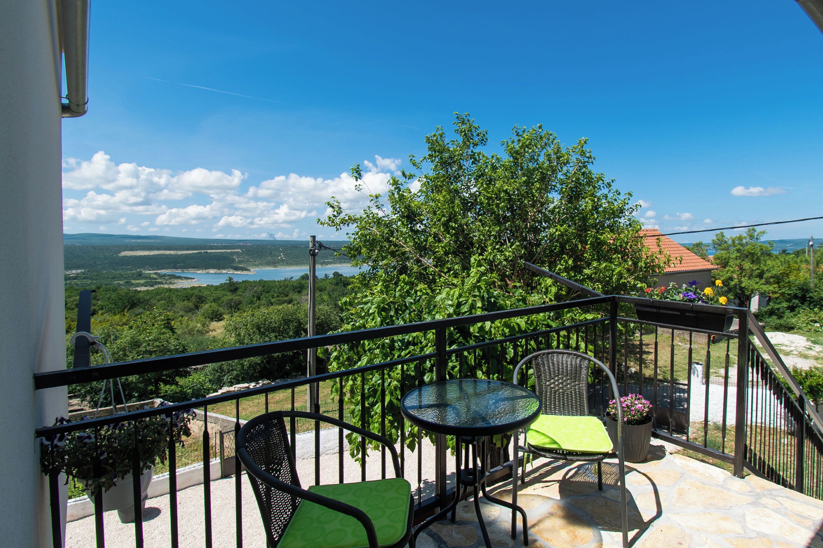 A balcony with a table and chairs, overlooking a scenic landscape with trees and a distant view of the sea.