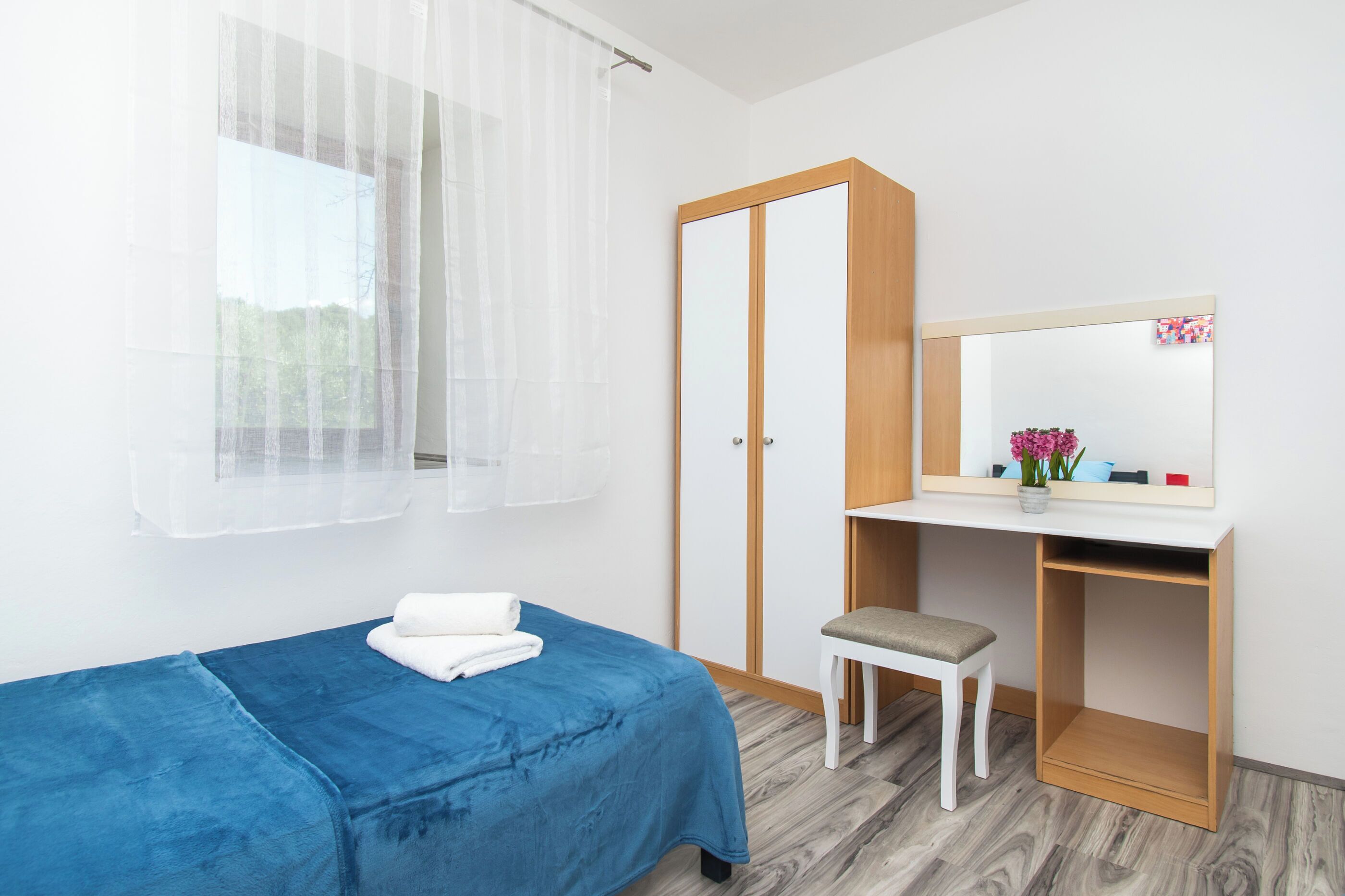 Une chambre à coucher comprenant un lit, un petit bureau, une chaise, une armoire et une fenêtre avec des rideaux légers.