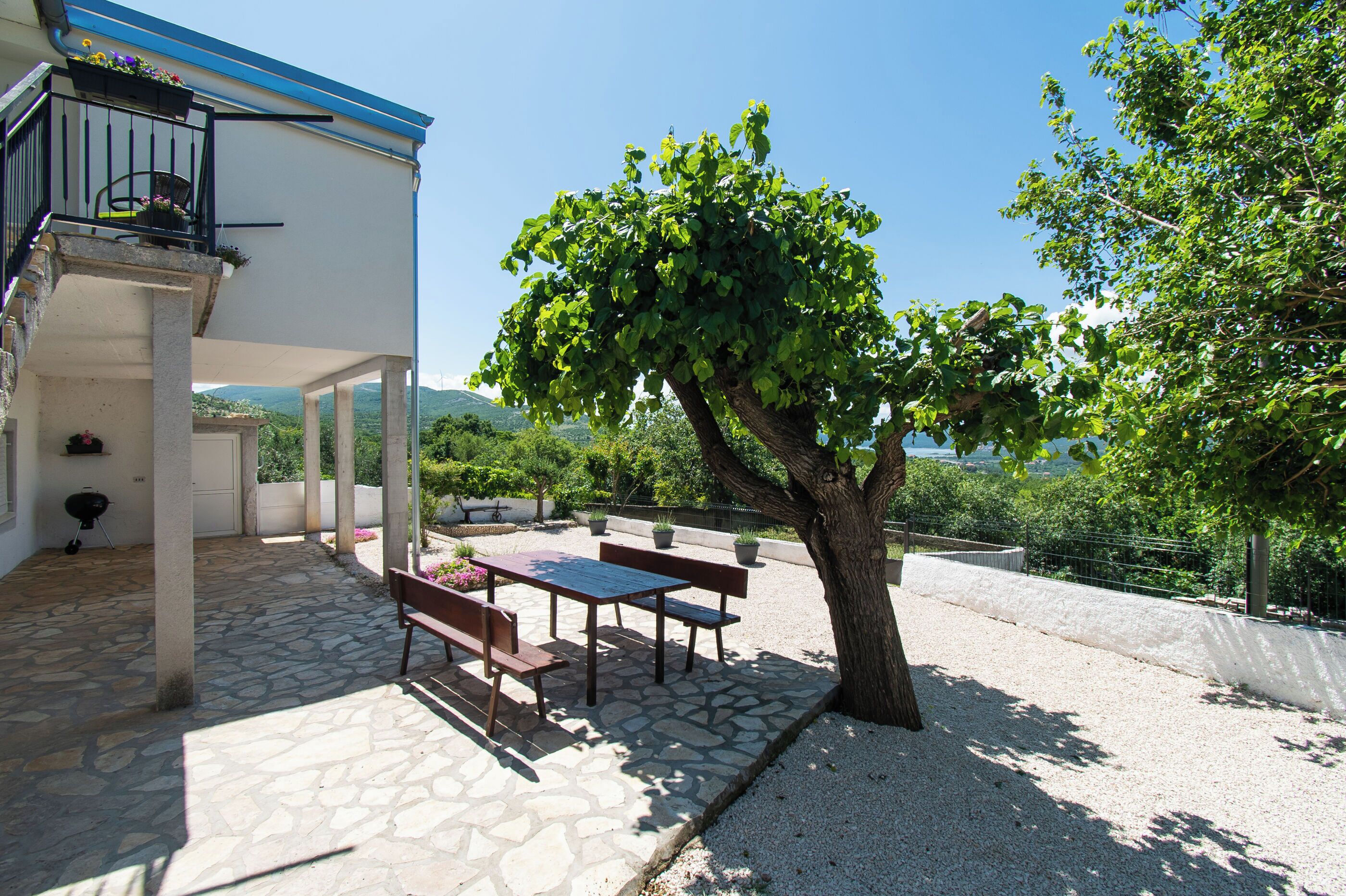 Une terrasse extérieure couverte, dotée d’un sol en pierre, d’une table et de bancs en bois, d’un arbre et offrant une vue sur le paysage environnant.