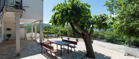 Une terrasse extérieure couverte, dotée d’un sol en pierre, d’une table et de bancs en bois, d’un arbre et offrant une vue sur le paysage environnant.
