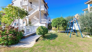 A two-story building with a white exterior, a balcony on the second floor, and a small garden area with a swing set.