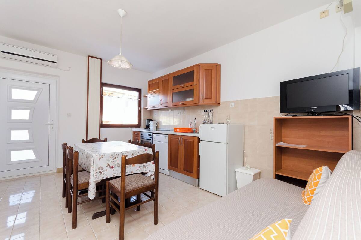 Une cuisine compacte avec des meubles de rangement en bois, un réfrigérateur, une table à manger avec des chaises et un téléviseur posé sur un meuble en bois.