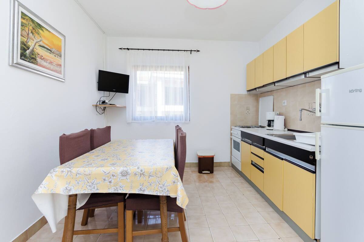 Une cuisine de taille réduite avec des meubles de couleur jaune, un réfrigérateur blanc et une table à manger recouverte d’une nappe à motifs floraux.